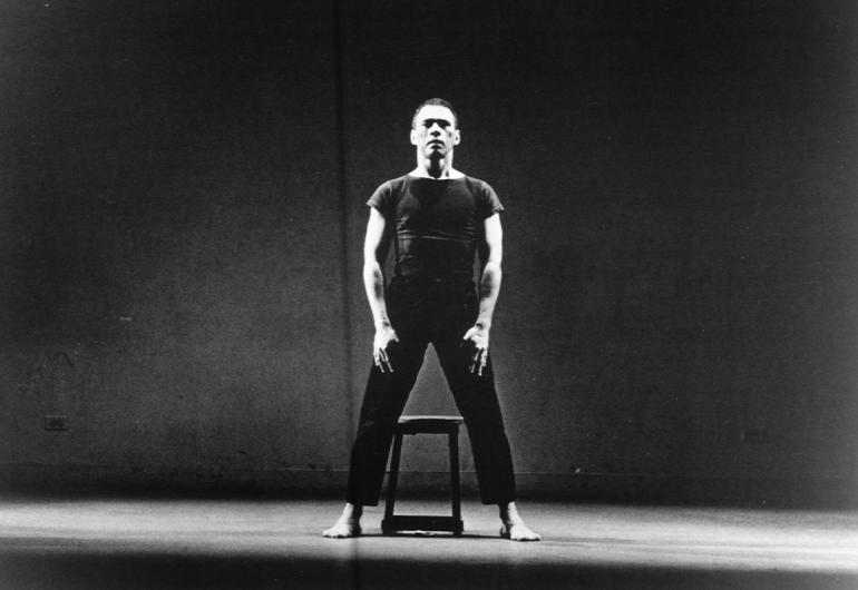 Victor Quijada stands firmly over a stool, legs apart and arms down, in spotlight against black background. 