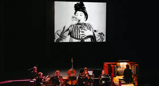 An ensemble of 7 musicians dressed in black perform onstage with a projection of a film above them. The screen shows a black and white film still of a person wearing a striped top, drinking a glass of wine with one hand held to their chest and their eyebrows raised.