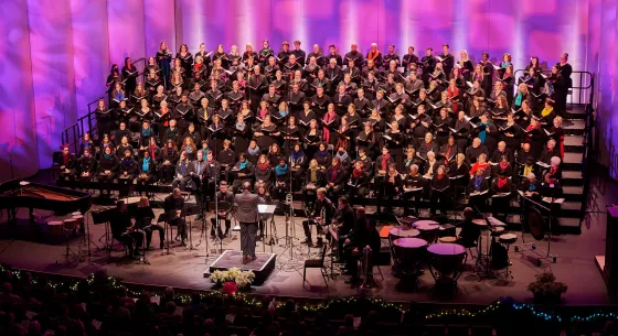 A choir wearing black with various colored scarves performs onstage with patterned pink and purple lighting projected behind them. A conductor stands downstage with their back toward the camera and musicians surrounding them.