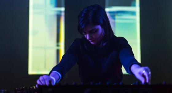 Image of Sarah Davachi in dark clothes and purple light with light pastel colors behind her using a soundboard.