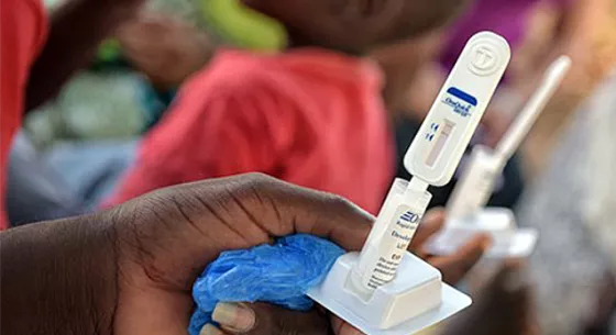 A dark-skinned hand holds up a small white medical device. Out of focus people are shown in the background.