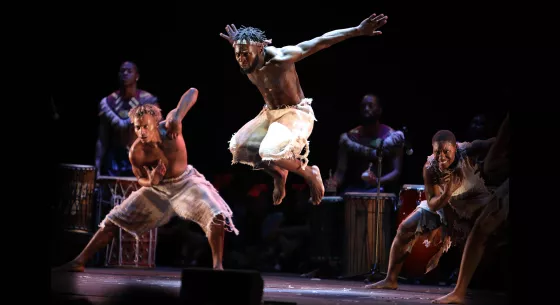 Three dancers wearing white appear on stage in front of musicians playing percussion. One dancer appears in the center, jumping in the air with their arms outstretched behind them.