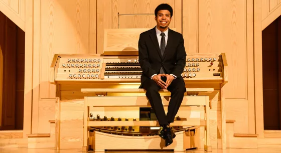 An organist with dark hair wearing a black suit and tie sits in a recital hall with their back towards a wooden organ console and fingers intertwined and placed on their knees.