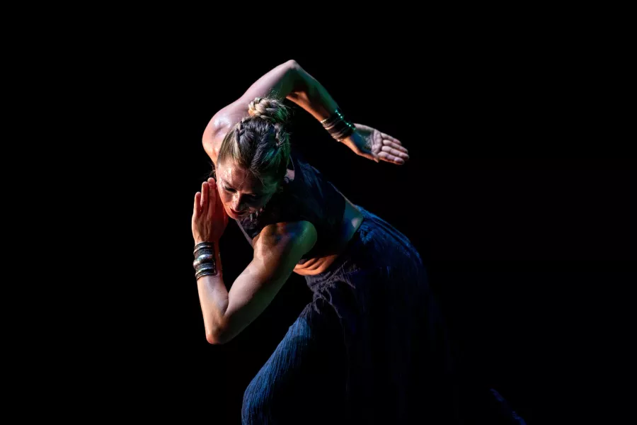 A dancer stands while leaning forward with their head down. One arm is bent behind their neck, while the other is positioned in bet post across their chest.