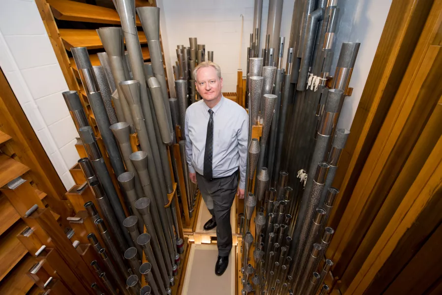 A person wearing a blue button-up shirt with a black tie is standing in between metal rods of various heights. There are also wooden panels around the walls.