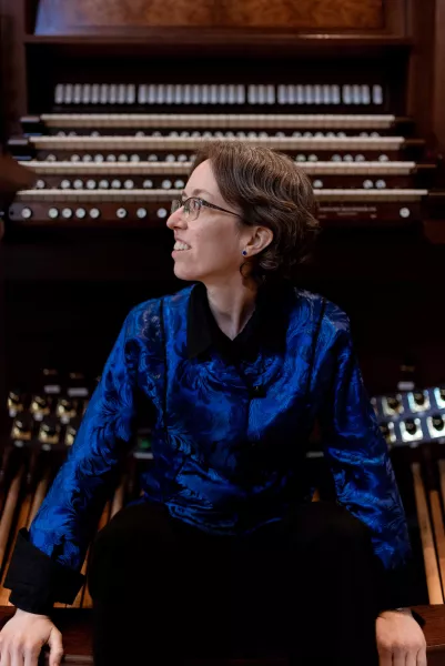 An organist wearing a deep blue button down with short, dark brown hair and glasses sits on a bench in front of a large, wooden organ console with their head turned to the right.