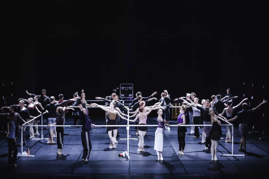 A large stage is outlined by a hollow square of connecting ballet barres. Dancers outline and interior and exterior of the square, all posed in fifth position.