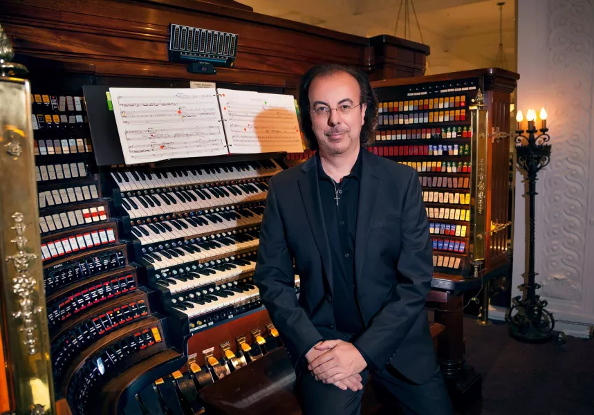 A black-haired person wearing glasses and all black sits on a bench with their back to an organ console with their hands crossed on their lap.