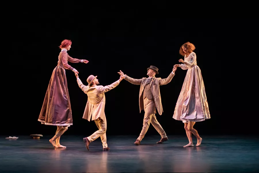 Two very tall people wearing dresses hold hands with a person wearing a hat. Each pair is facing one another and appear to be walking towards each other.