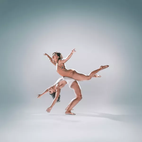 A dancer wearing white is standing arching their back with a dancer wearing beige balanced on their stomach.