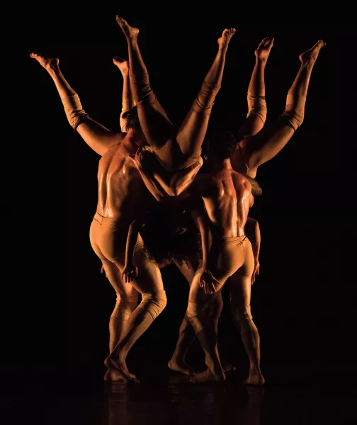 On a dimly lit stage, three dancers are held upside down in a circle while three other dancers hold them.