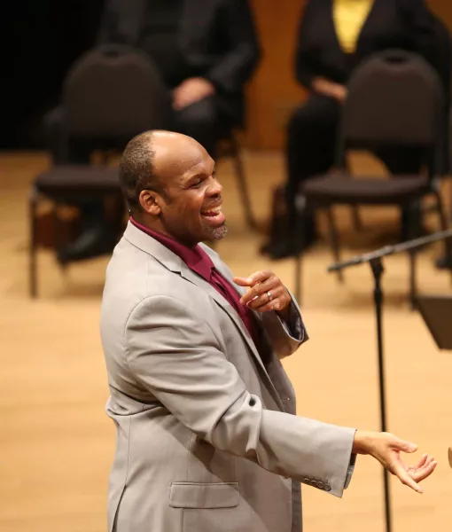 A man wearing a light grey suit with a dark red collared shirt, smiling and making an arm gesture.