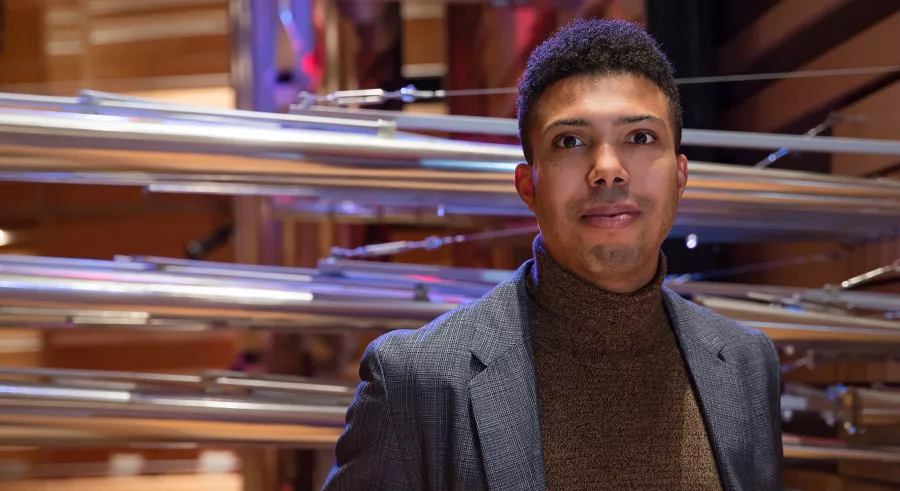 A person with dark hair wearing a brown turtle neck under a dark gray suit coat stands toward the camera with large organ pipes behind them.