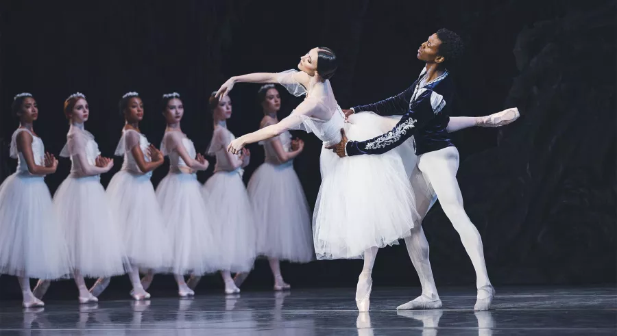 A dancer (man) steadies a ballerina as she reaches forward on pointe.