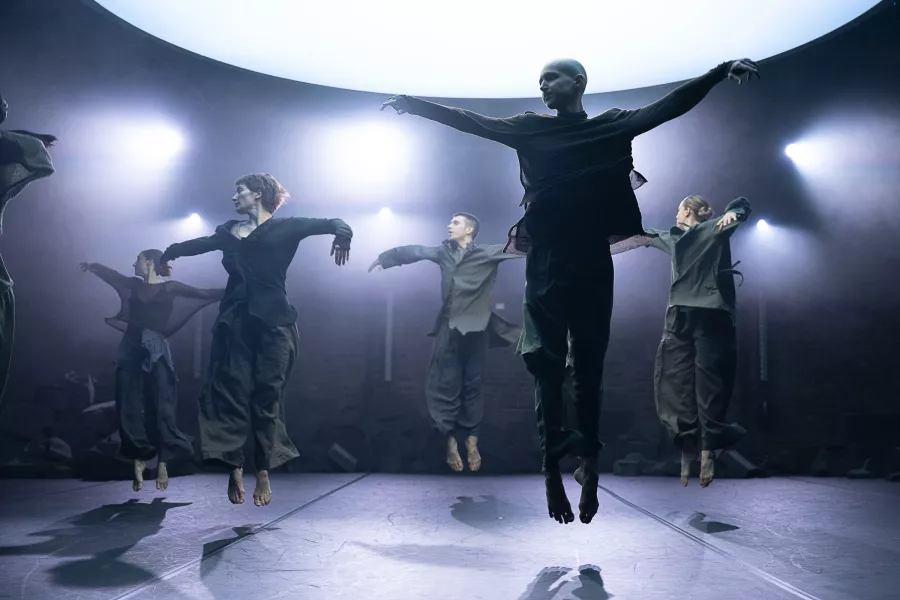 Group of dancers looking to their right, jump straight up with toes pointed, arms out to the sides. They are on a stage with many hazy spotlights behind them creating a dramatic lighting effect.