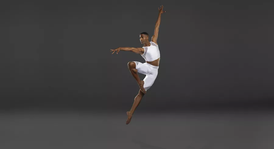 Male dancer in white leaps with toe to opposite knee and arms up and out