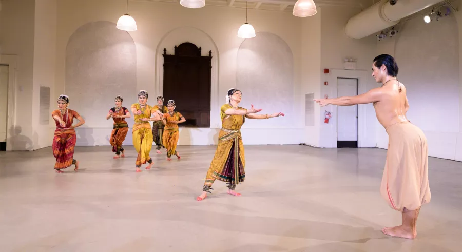 Dancer in white reaches toward a dancer in yellow garb. Other performers run stage right.