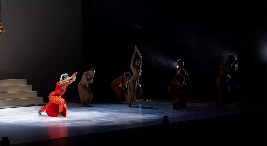 A dancer in red lunges in spotlight, others move in shadow.