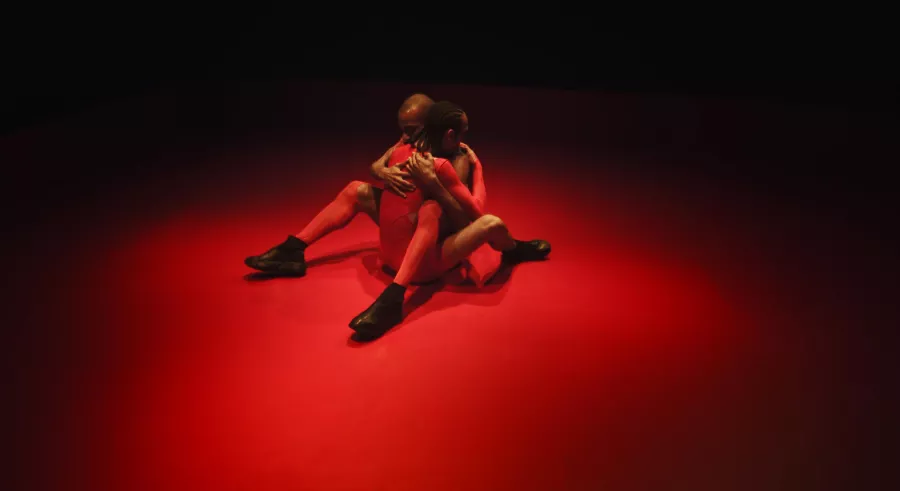 Dancers intertwine legs on red stage floor.