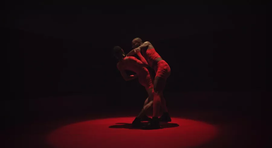 One dancer supports the other's frame. They are cast in half-dark red lighting.