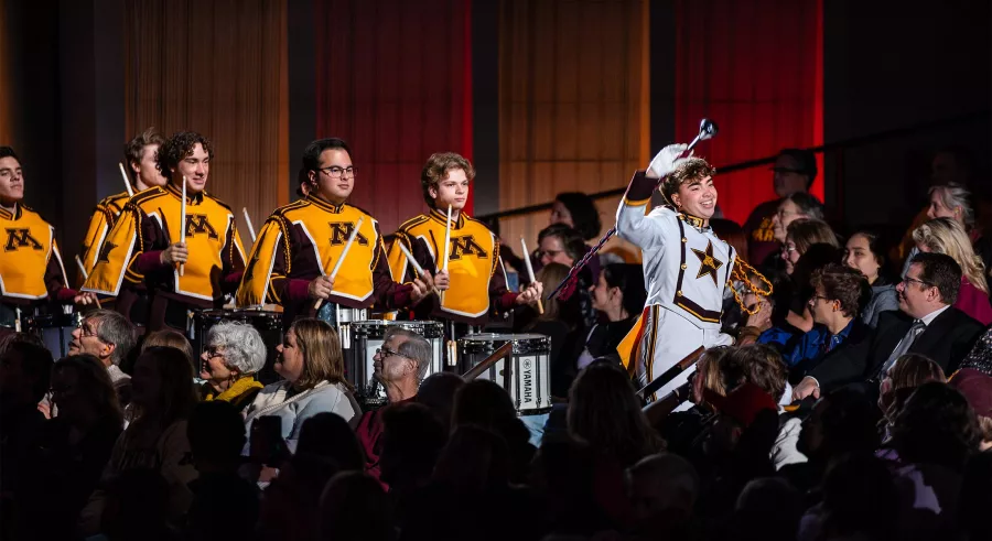 Uniformed conductor and drummers march through a crowd.