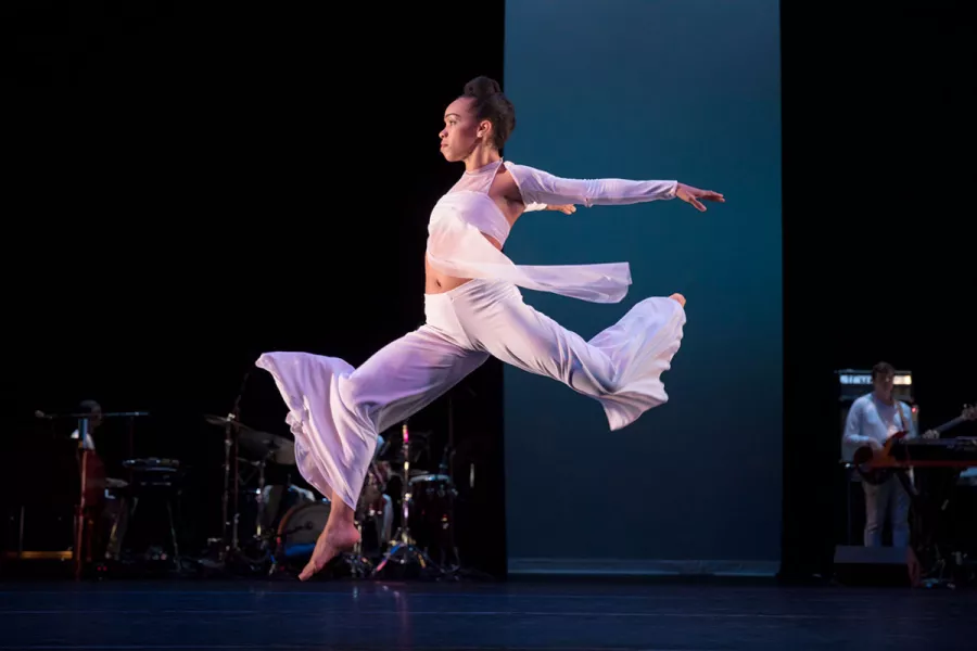 A female dancer wearing white leaps forward on stage.