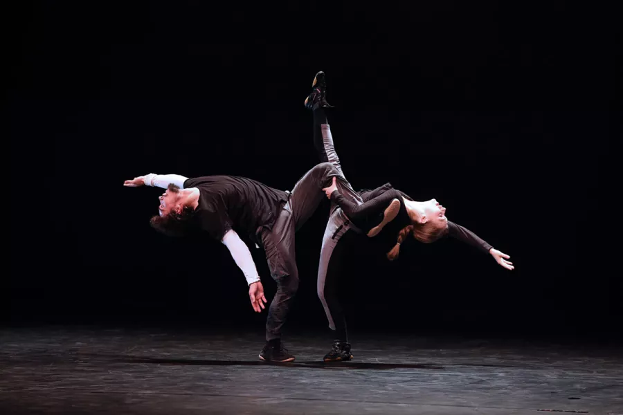 Two dancers stand facing eachother on one foot kicking while arching backwards in opposite directions