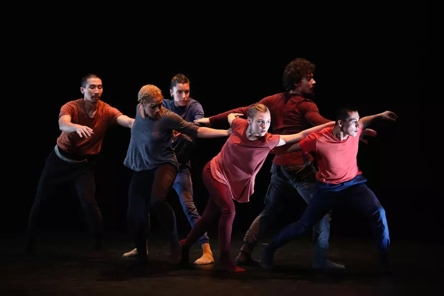 Six dancers in colorful tops, hands on the others shoulders in line with two in the middle pull the group forward