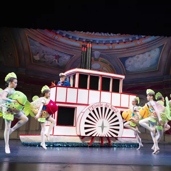 Young dancers wearing fish costumes dance in front of a large paddleboat prob