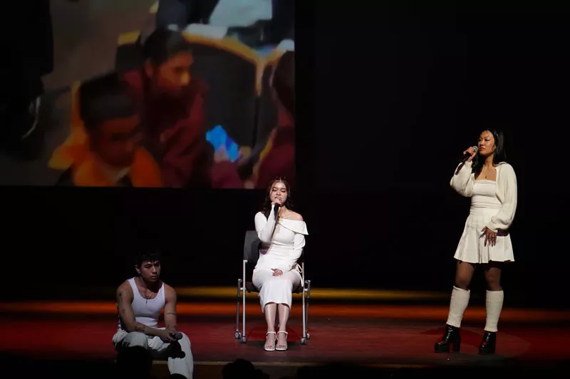 Three women wearing different white outfits sing on stage