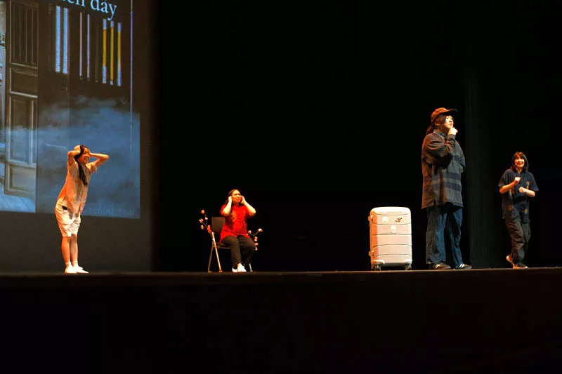 Four performers perform a skit on stage. A suitcase is a prop.