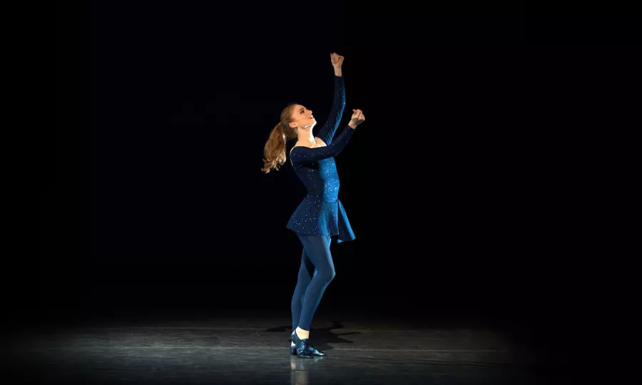 Female in dark blue stands with one heel raised and knee bent, looking up with arms up like she's snapping