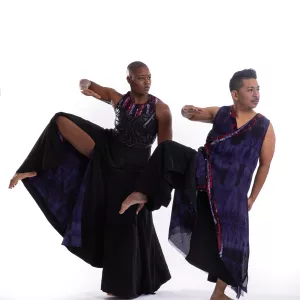 Two dancers in black against a white background, stand on one foot, right arm lifted and bent at elbow, right knee raised to elbow,.