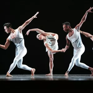 Dance Theatre of Harlem dancers wearing white perform on stage with a black background.