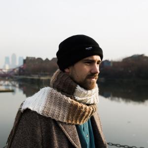 Kit Downes wearing a knit scarf and hat, stands outside by a lake with cityscape in distance.